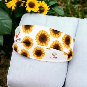 Sunflower FND awareness headband folded and displayed outdoors on a grey surface with green leaves and yellow sunflowers in the background.